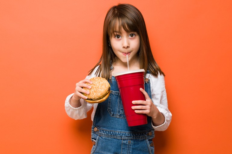 Portrait of a little girl dressed casually and taking a sip of a large soda while holding a burger in the other hand; Shutterstock ID 2151585909; purchase_order: aj; job: ; client: ; other: