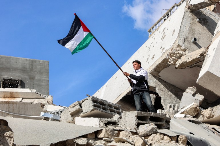 A man holds up a Palestinian flag while standing atop the rubble of a collapsed building at the Jabalia camp for Palestinian refugees in the northern Gaza Strip on January 19, 2025 after a ceasefire deal in the war between Israel and Hamas was implemented. The long-awaited ceasefire in the Israel-Hamas war was delayed January 19 after Prime Minister Benjamin Netanyahu said at the last minute that it would not take effect until the Palestinian militant group provided a list of the hostages to be released. (Photo by Omar AL-QATTAA / AFP)