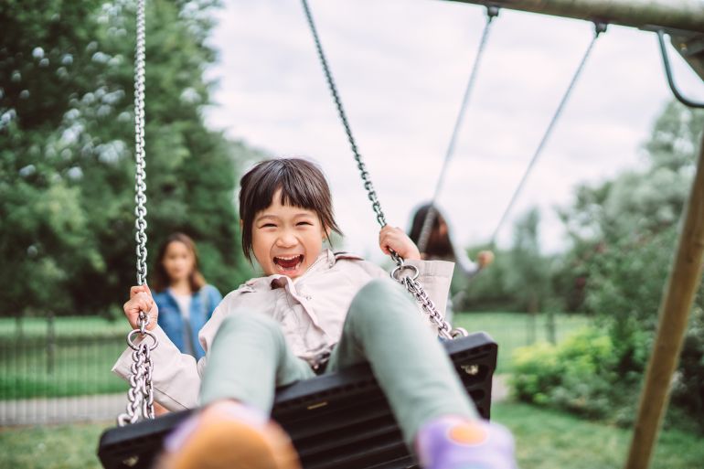 Lovely little girl smiling at the camera while playing on a swing set in playground joyfully.